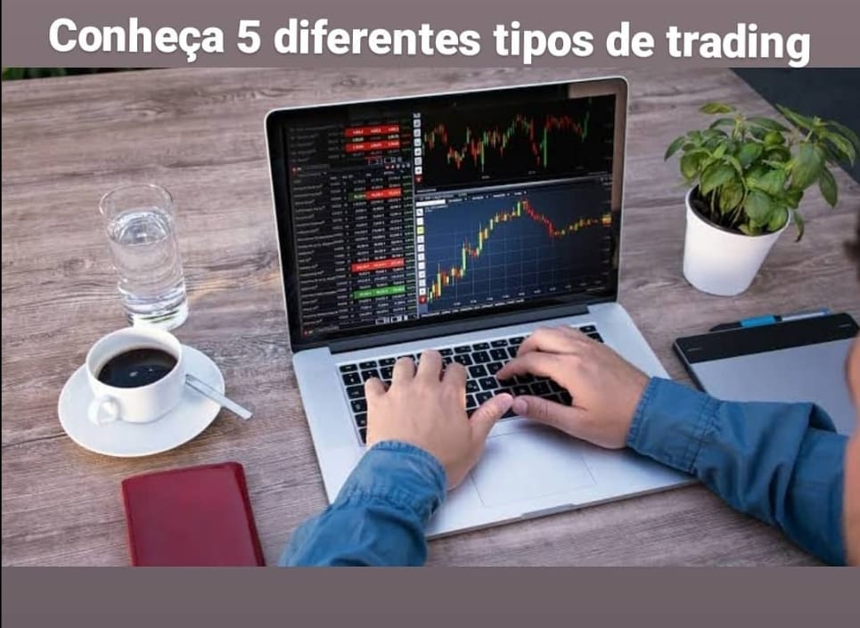 Imagem de uma pessoa trabalhando em um laptop com gráficos de trading na tela. O texto "Conheça 5 diferentes tipos de trading" está sobreposto na parte superior em letras brancas. A mesa de madeira contém um copo d'água, uma xícara de café com pires, um caderno vermelho, uma planta em um vaso branco e uma caneta. A pessoa usa uma camisa jeans azul e está digitando no laptop.